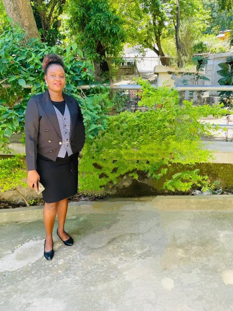 Femme en tenue professionnelle noire et gilet rayé posant devant une végétation verdoyante.